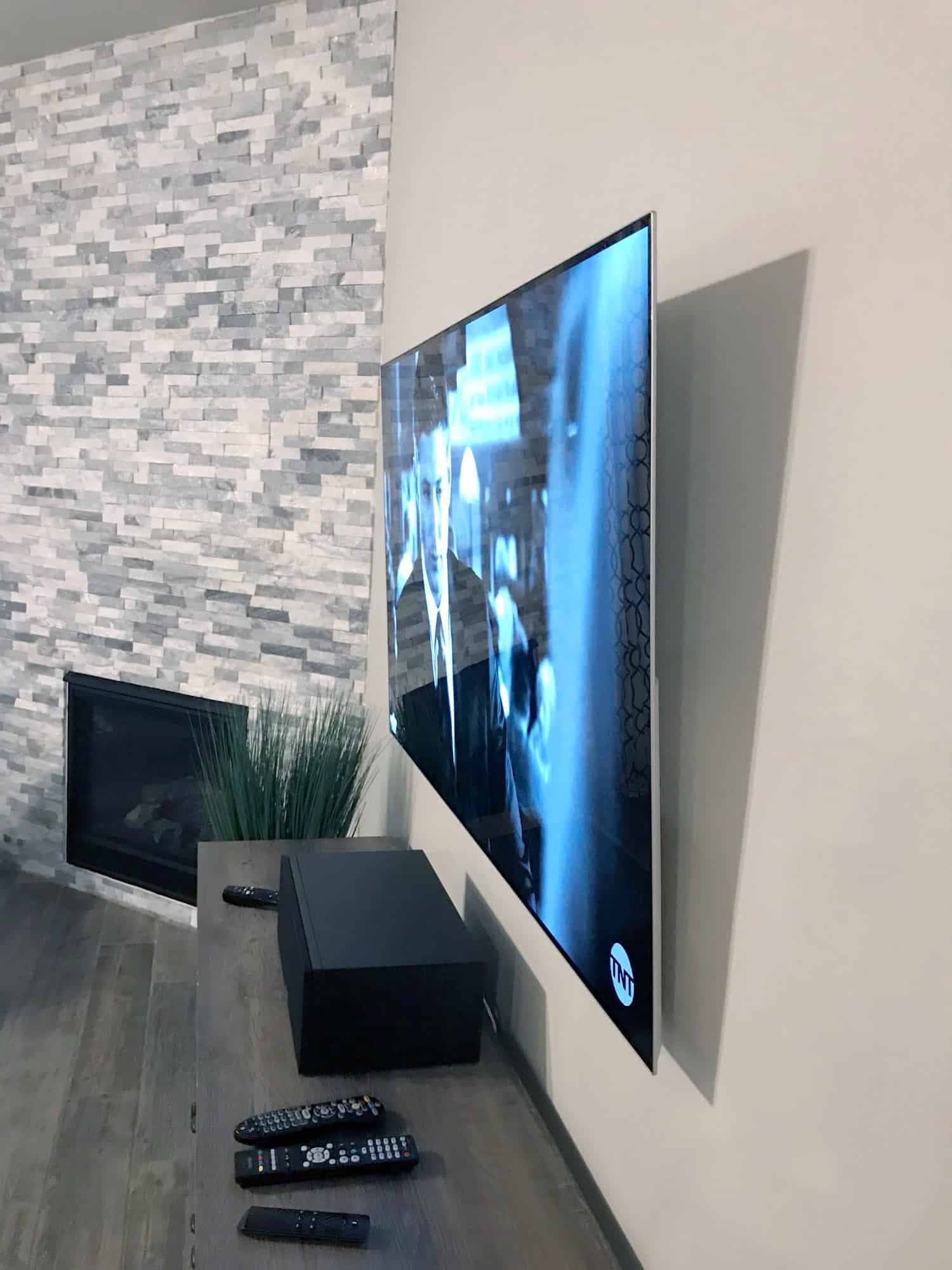 TV mounted at an angle beside a stacked-stone accent wall, showing a TNT drama above a sound base speaker