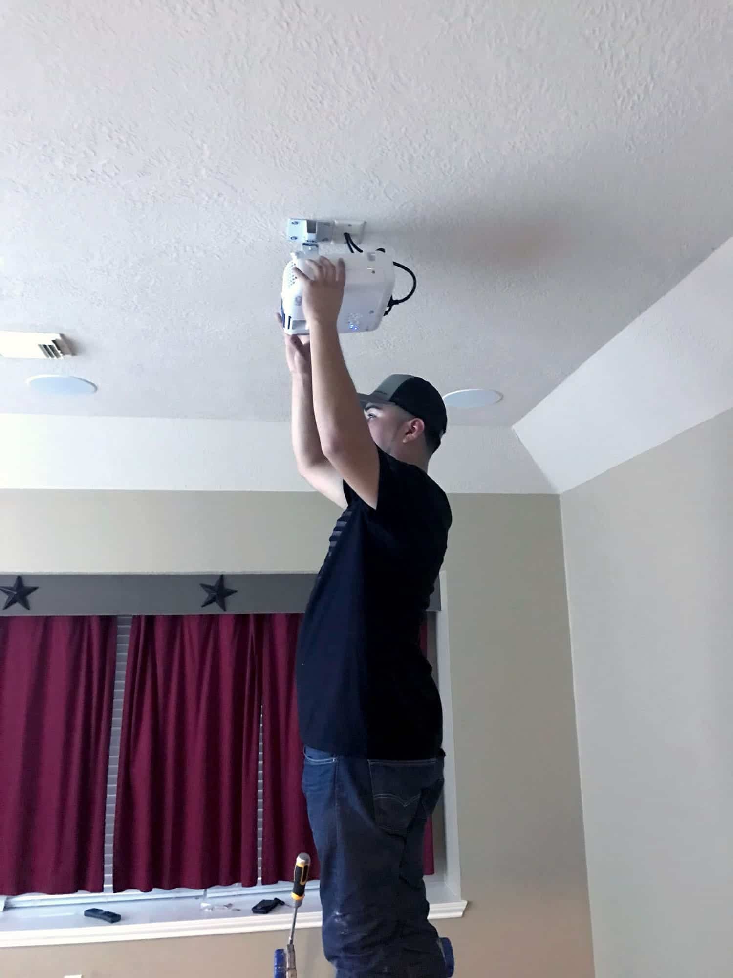 Installer on a ladder wiring an in-ceiling projector mount in a media room with patterned curtains