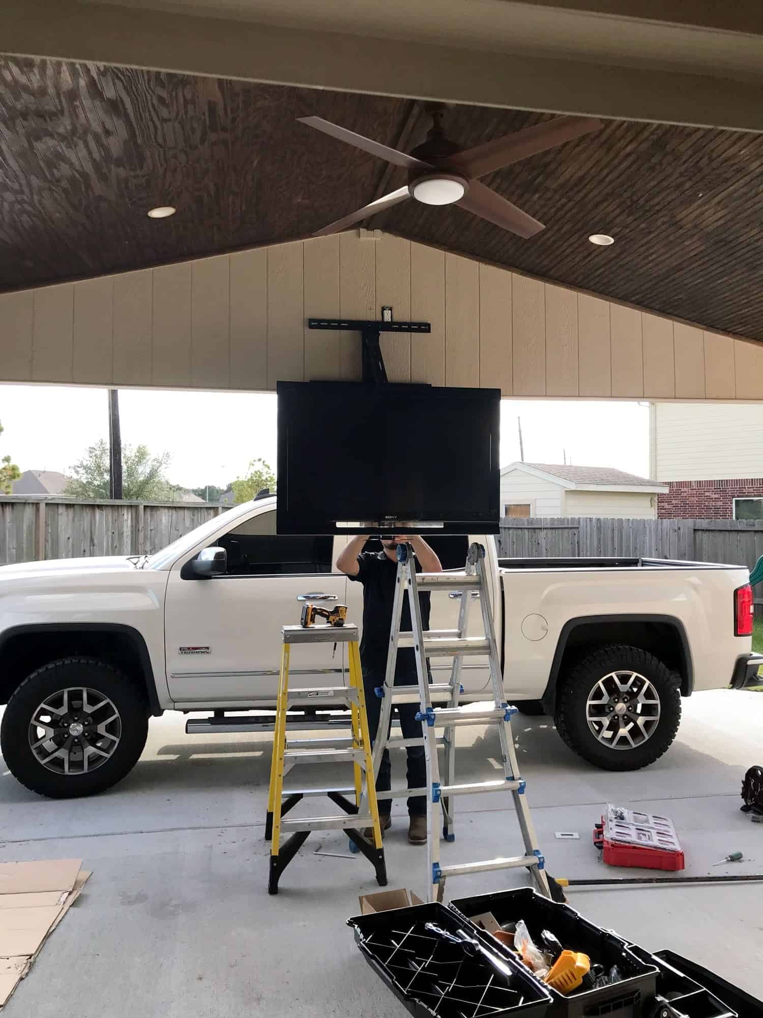 Two installers on ladders mounting a TV on a motorized lift above a pickup truck on a covered patio