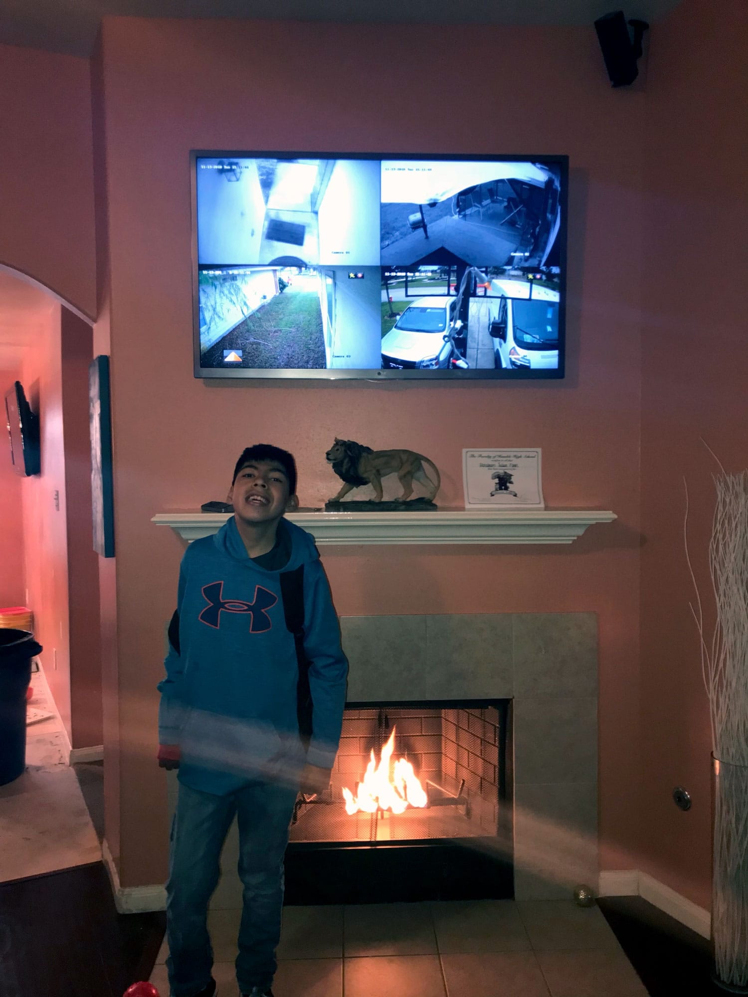 Security monitor above a fireplace showing four camera feeds, with a boy standing near the lit fireplace