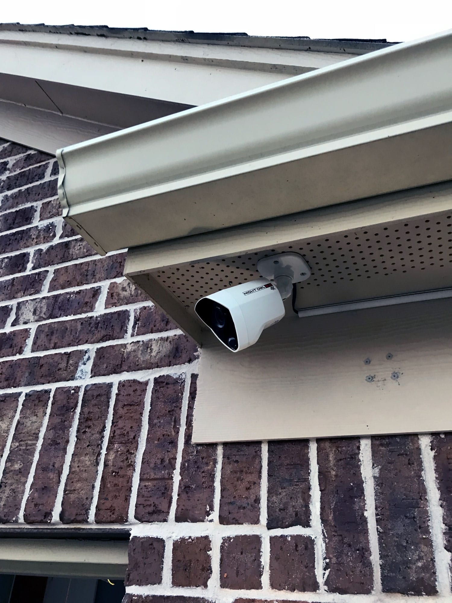 Close-up of a white outdoor security camera mounted under a brick home's eave