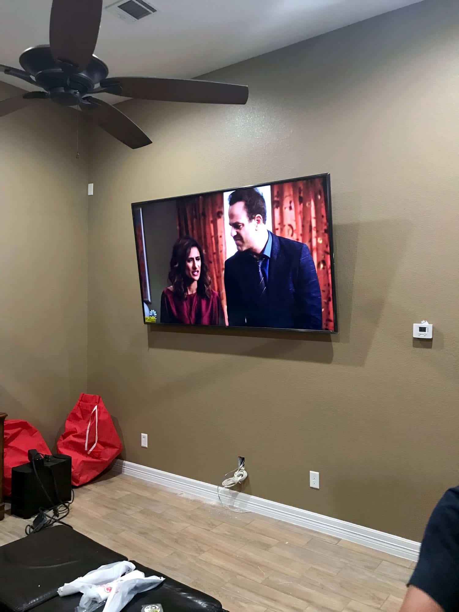 Large flat screen TV mounted on a living room wall below a ceiling fan, with maroon curtains in the background