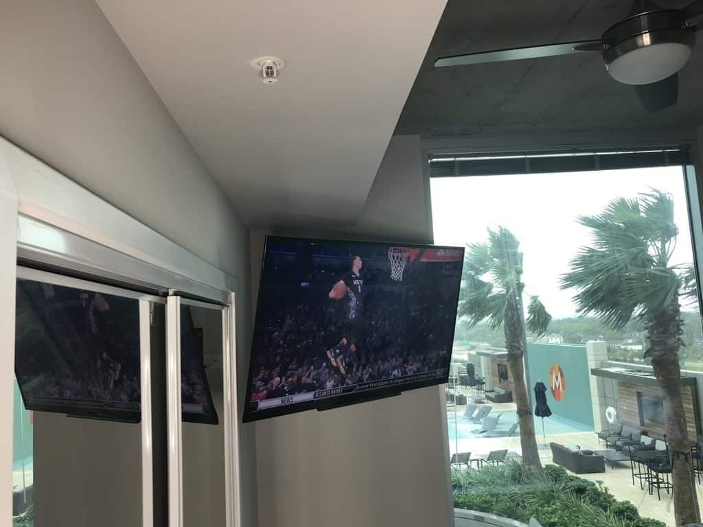 TV mounted in the corner of a covered clubhouse balcony overlooking a pool deck lined with palm trees