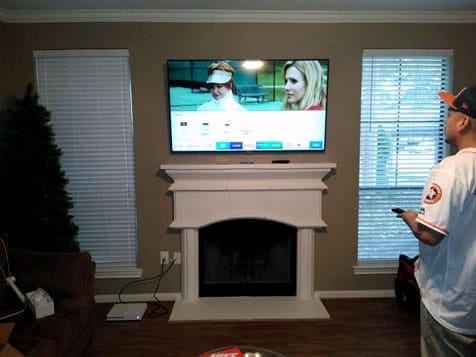 TV mounted above a white fireplace mantel showing a golf broadcast, with a man in a cap nearby