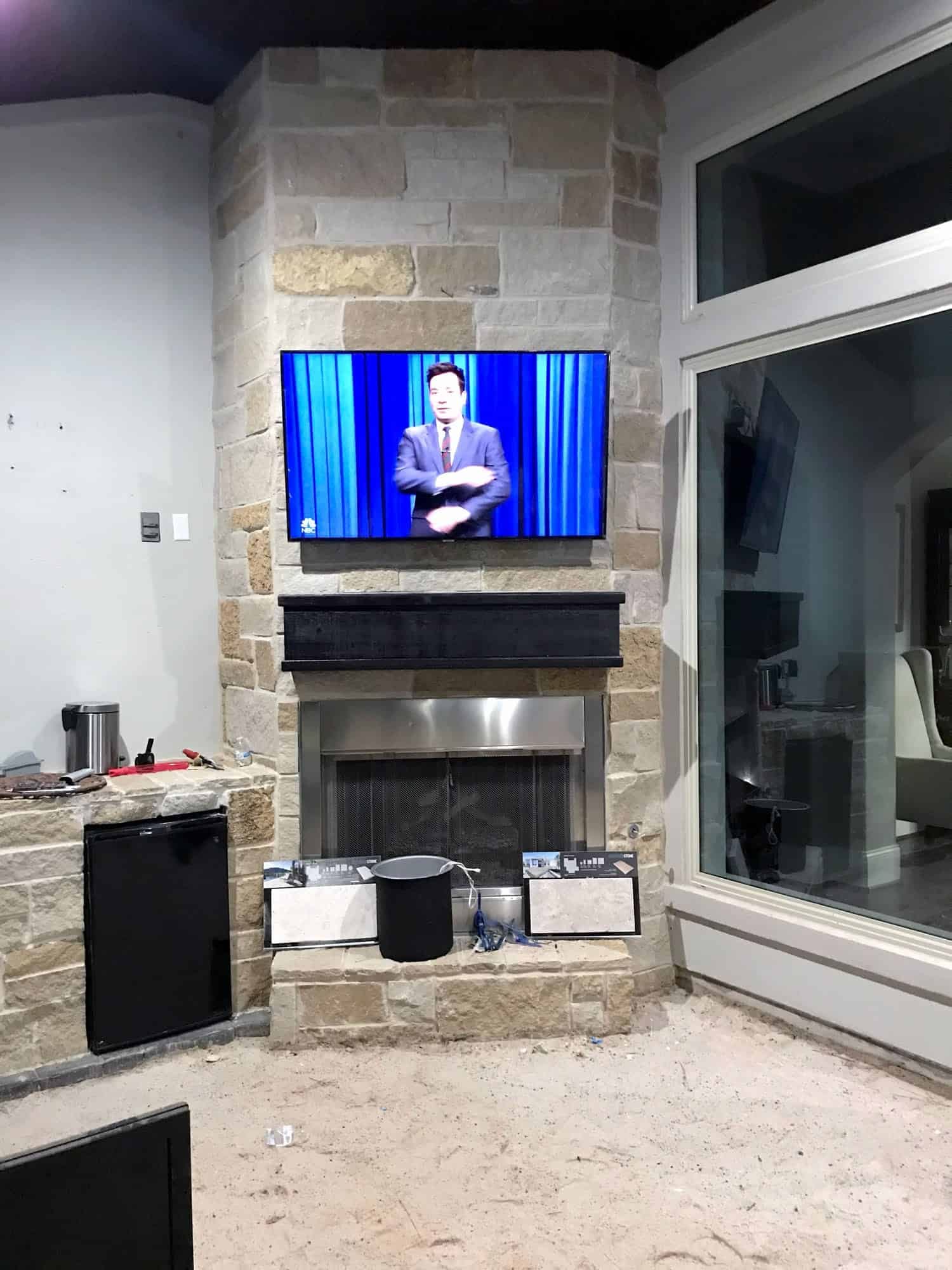 TV mounted on a stone fireplace surround showing a late-night talk show, with a mini fridge nearby