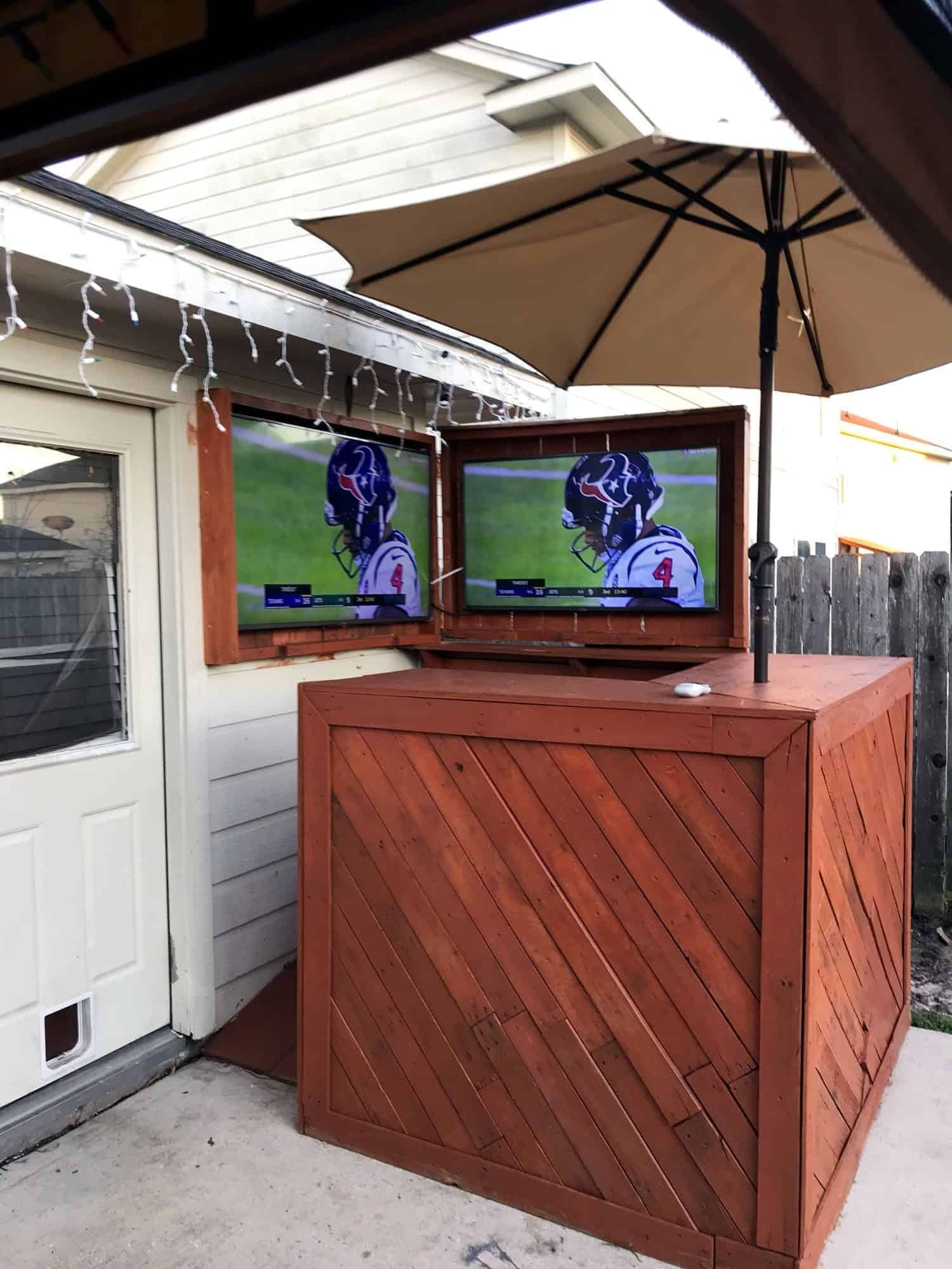 Two TVs mounted in wooden cabinet doors on an outdoor patio bar, both showing a football game