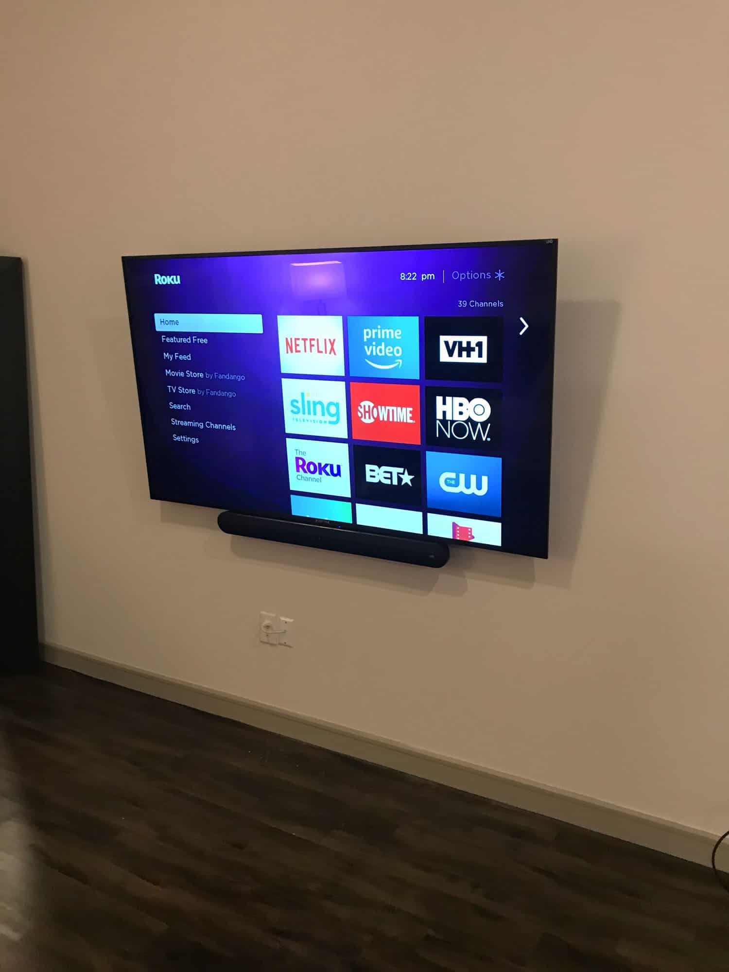 TV mounted on a wall with a soundbar below, displaying the Roku home screen with streaming app icons