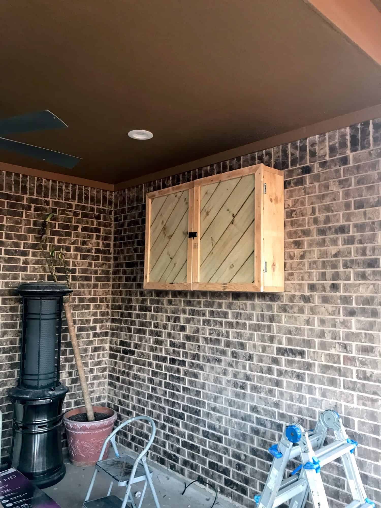 Closed wood cabinet with diagonal-plank doors mounted on a brick wall to conceal an outdoor TV