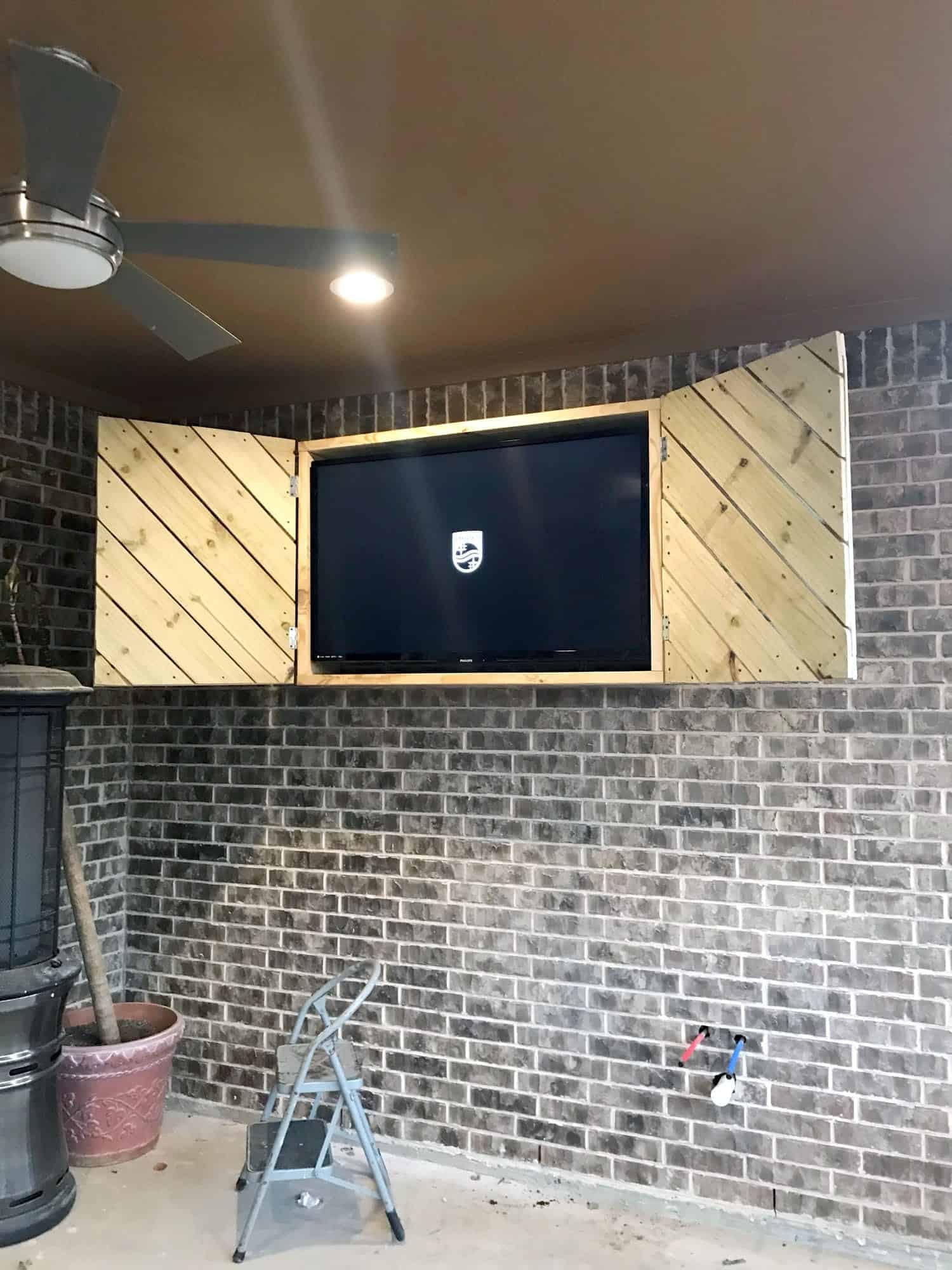 TV mounted in an outdoor brick alcove with wooden hinged doors, displaying a screensaver logo