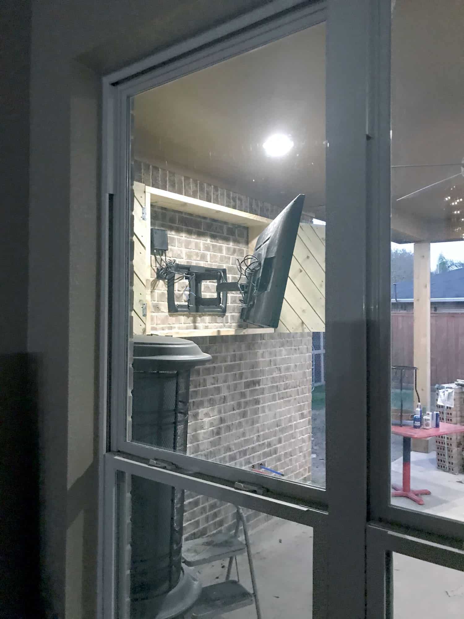 View through a patio door of a TV mounted in a partially built brick and wood enclosure with wiring exposed