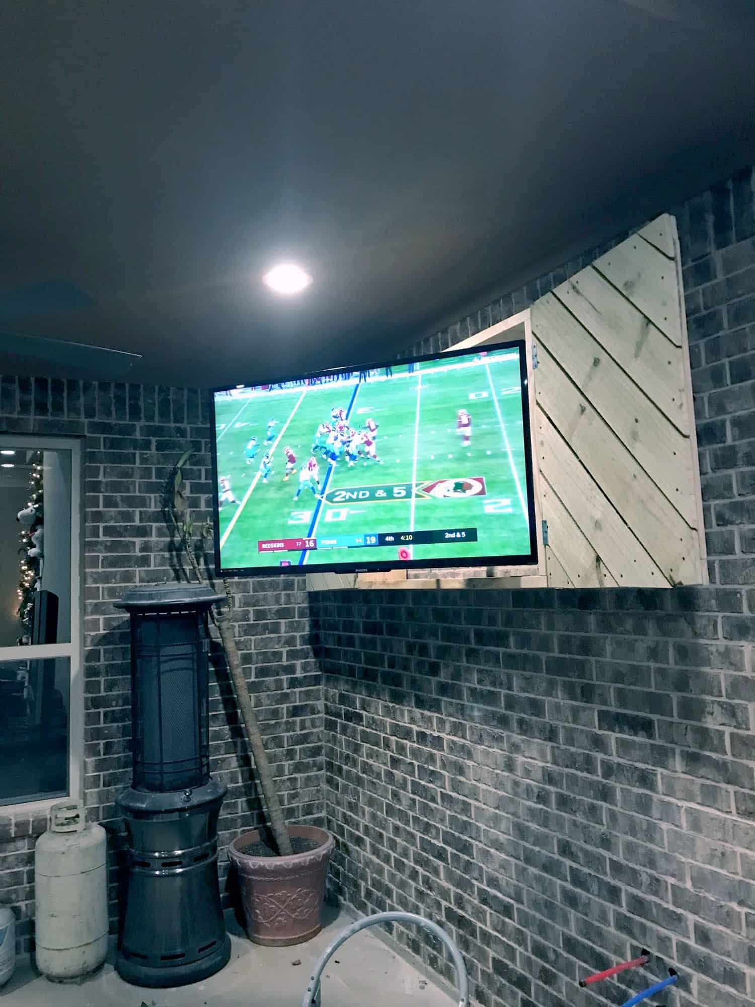 TV mounted in a brick outdoor wall cutout with a wood plank door, showing a football game