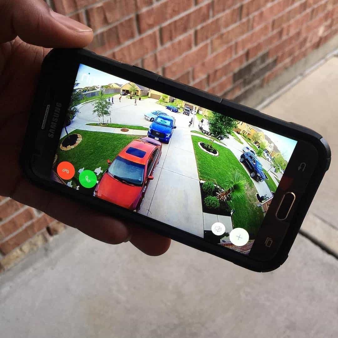 Hand holding a smartphone showing a live security camera view of a driveway and front yard