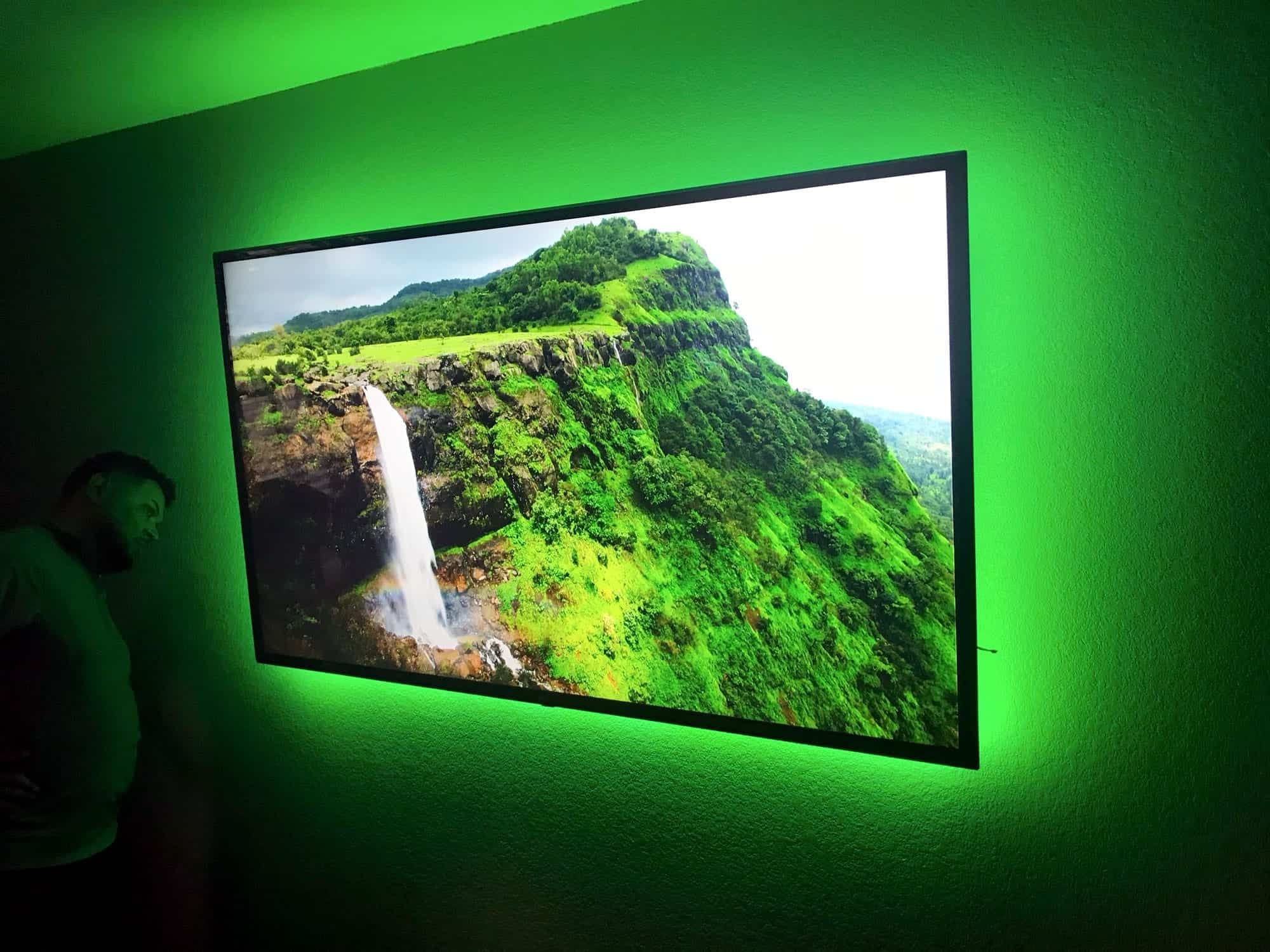 Wall-mounted TV with green LED backlighting displaying a waterfall and mountain scene in a home theater room