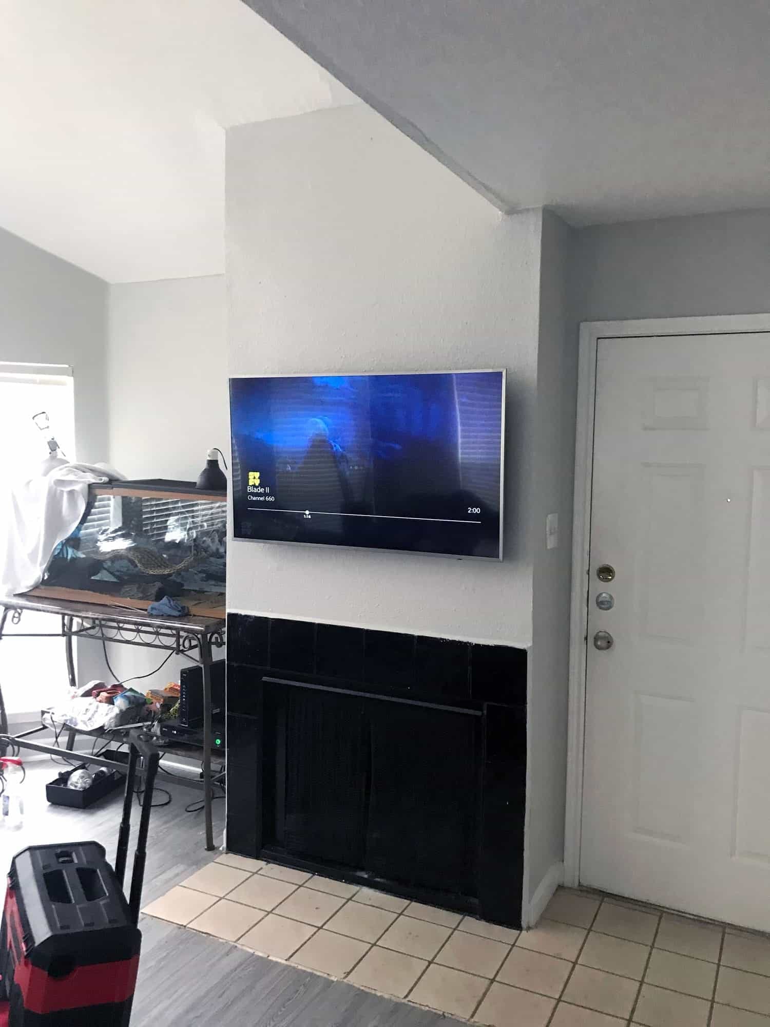 TV mounted above a black-tiled fireplace playing a movie, with a reptile terrarium in the room