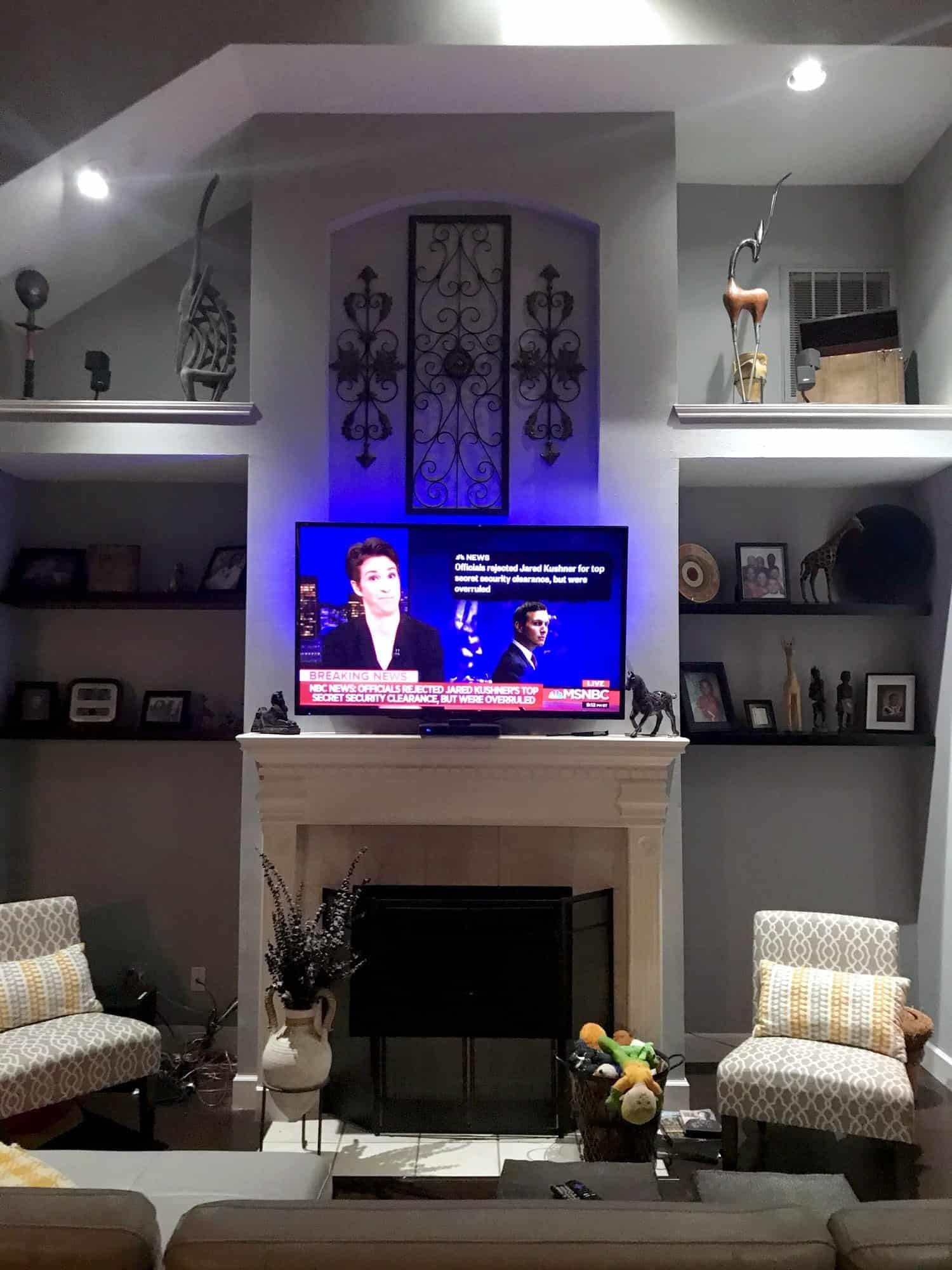TV mounted above a white fireplace showing a breaking news broadcast, framed by wrought iron wall art and built-in shelving