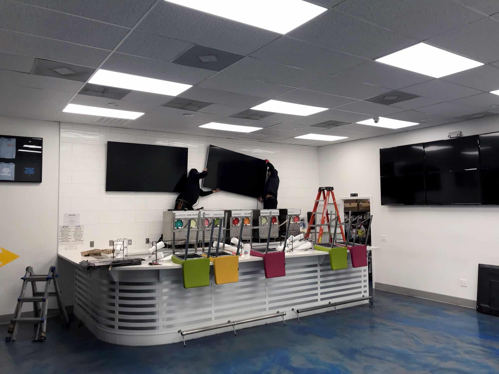 Two installers mounting dual TVs above frozen drink dispensers in a commercial bar space