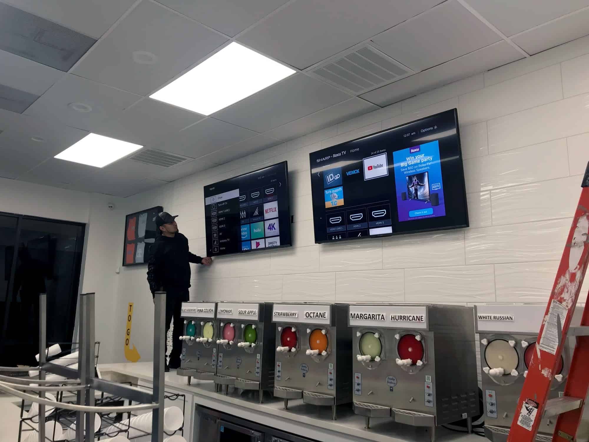 Installer standing on a counter mounting two TVs above a row of frozen drink dispensers in a commercial space