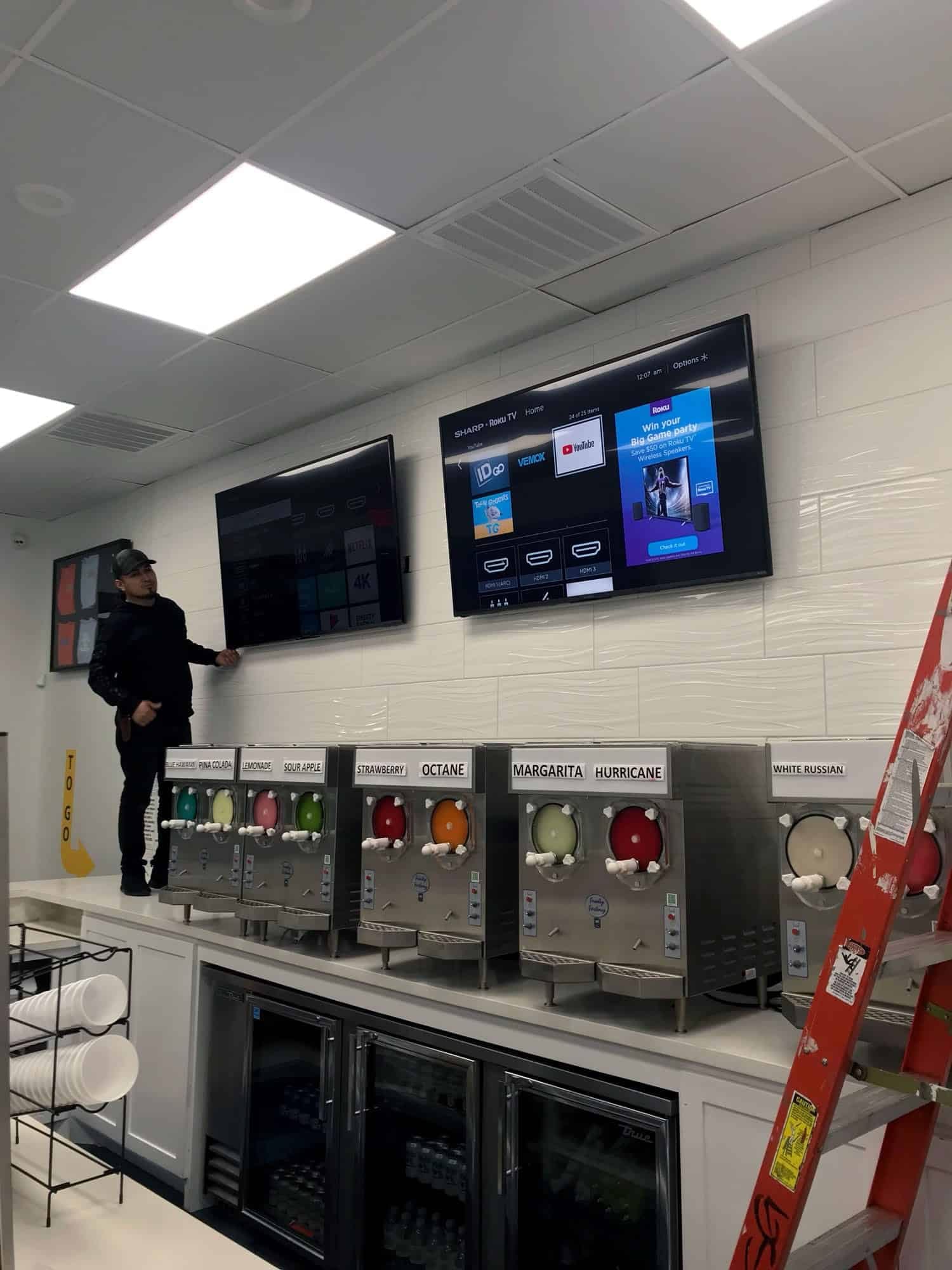 Two wall-mounted TVs displaying streaming menus above colorful frozen drink machines in a commercial space