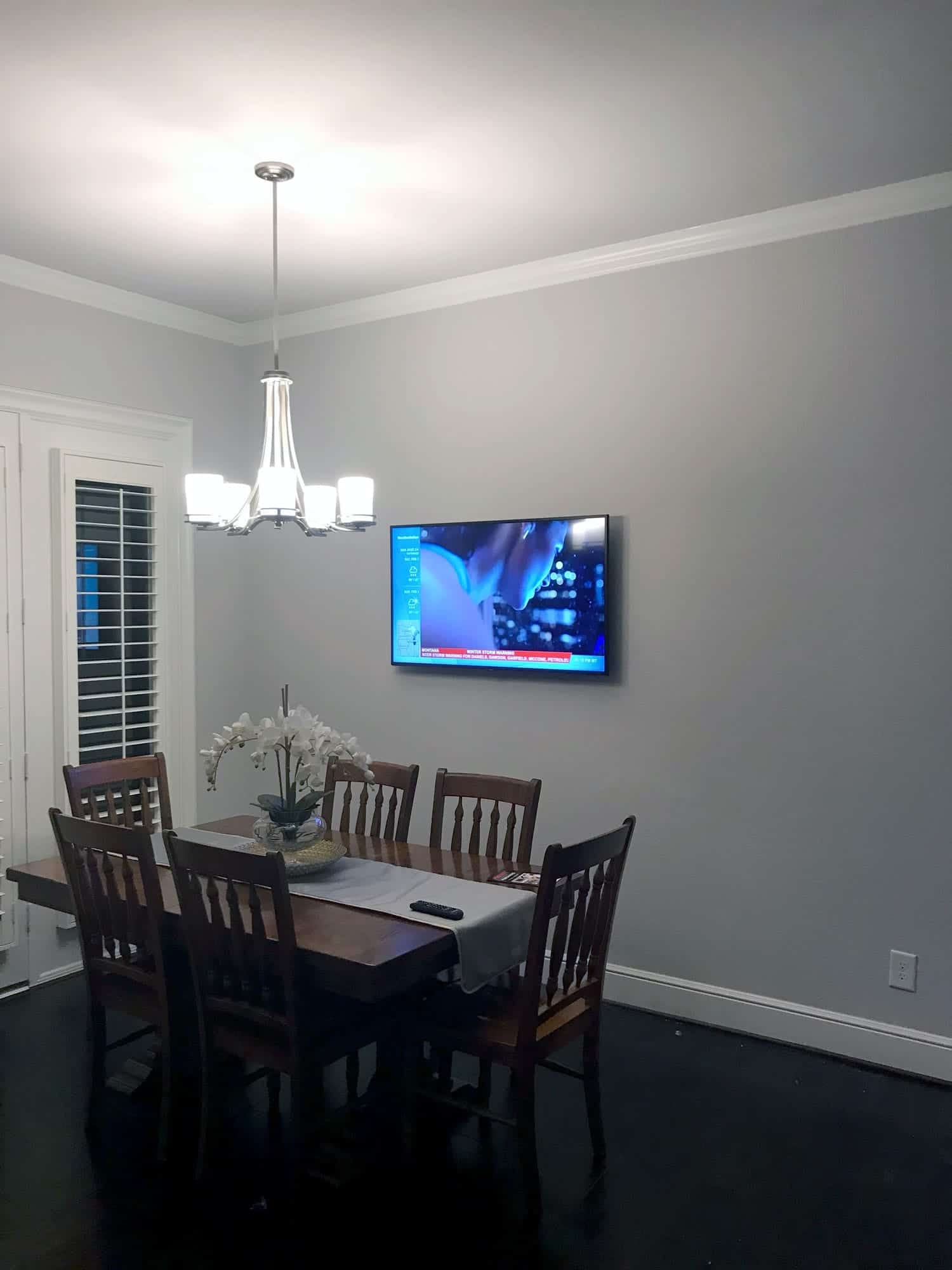 TV mounted on a dining room wall showing a weather broadcast beneath a crystal chandelier