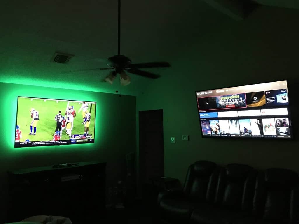 Two TVs mounted in a dark home theater room with green LED backlighting and theater seating