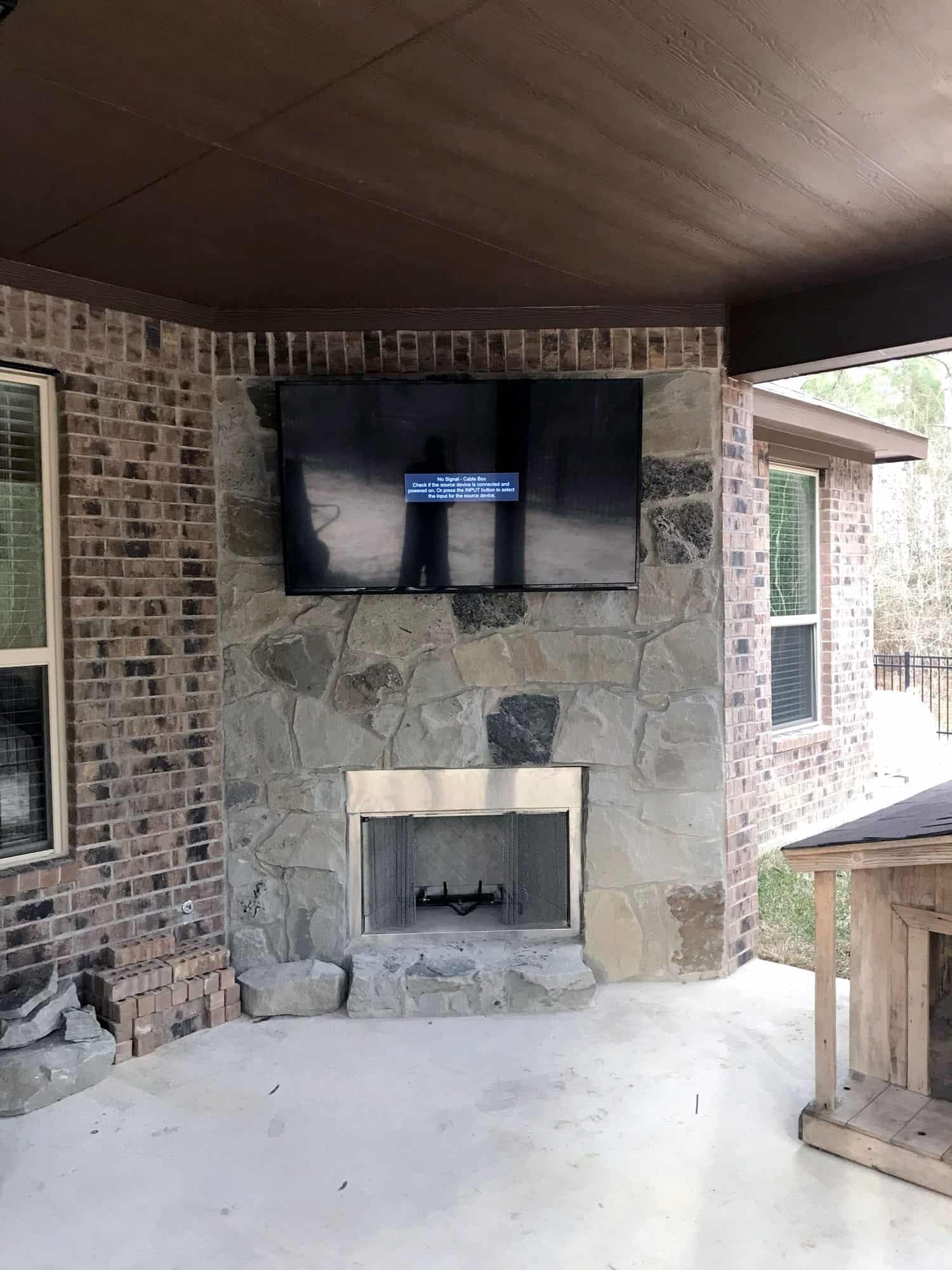 Wide view of a TV mounted on an outdoor stone fireplace column with a no-signal cable box message, flanked by brick walls and windows on both sides, with a small wood structure with a shingled roof on the covered patio