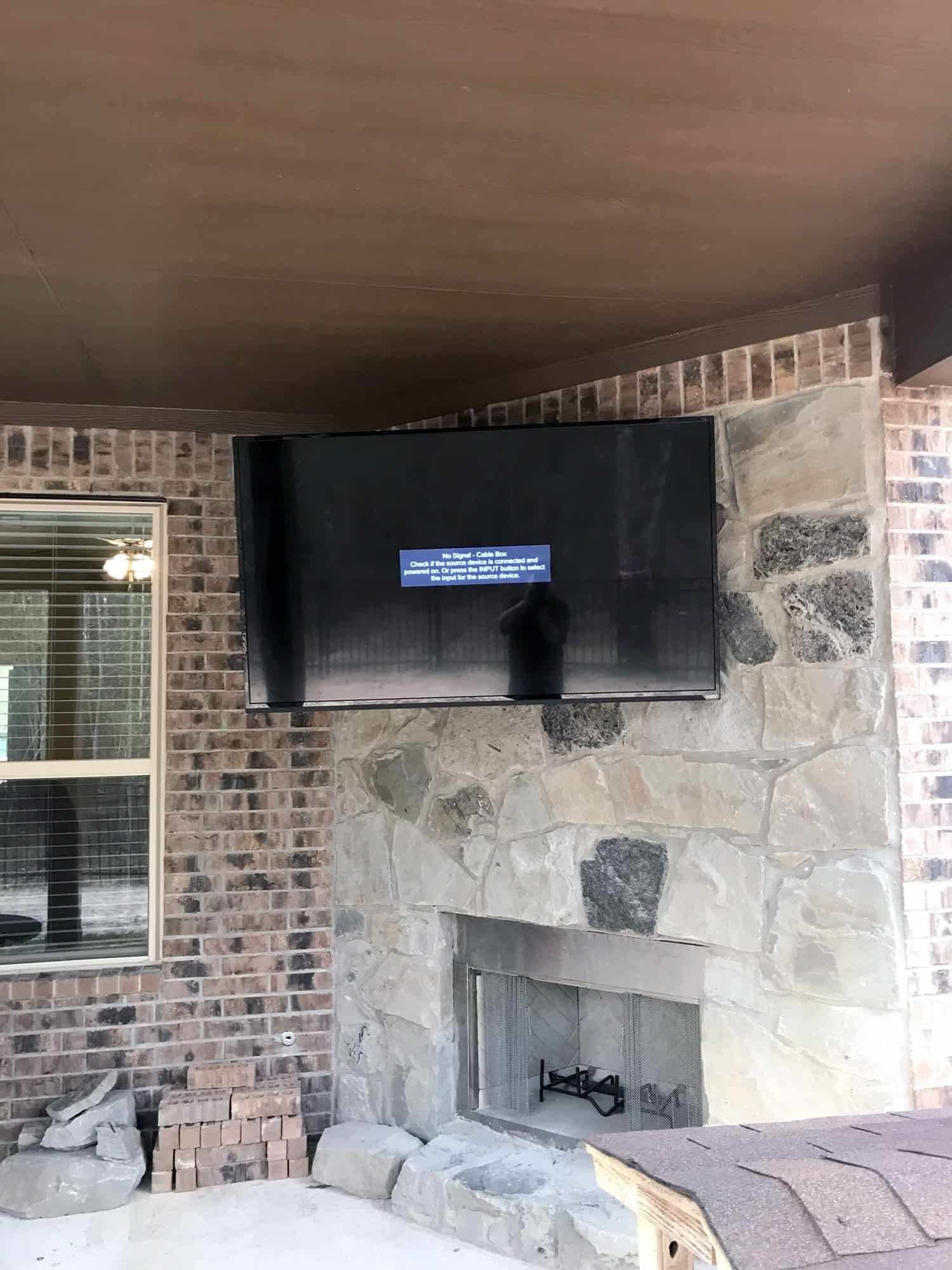 Closer view of the same outdoor stone fireplace TV showing a no-signal cable box message and the VIZIO logo on the bezel, with the house's brick corner and roof shingles visible nearby