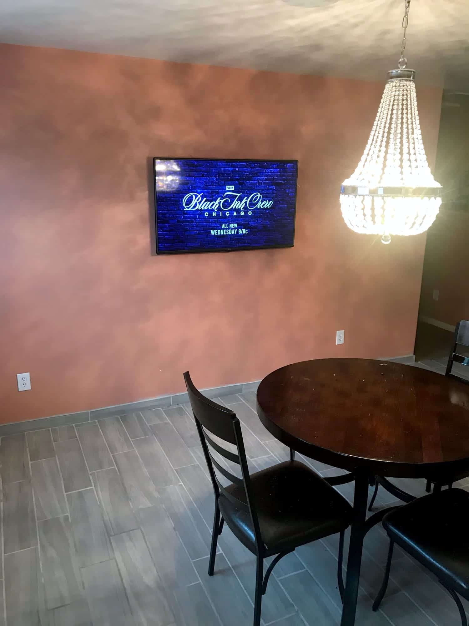 TV mounted on a terracotta accent wall showing a VH1 show promo, beneath a crystal chandelier