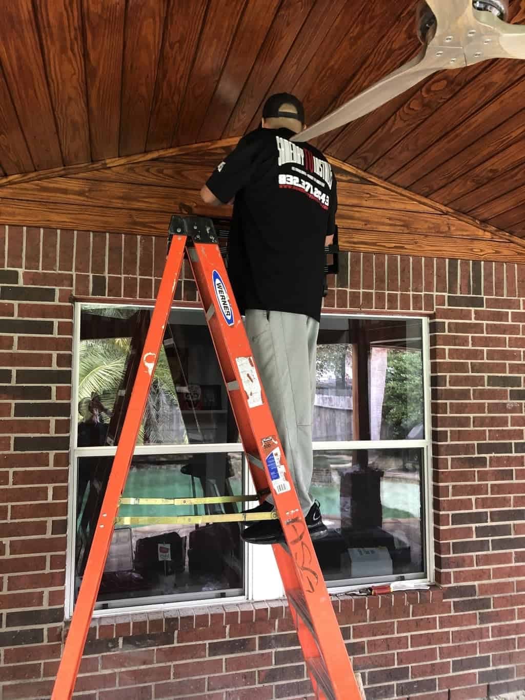 Installer in a branded Same Day TV Installs shirt on a ladder working under a covered patio roof